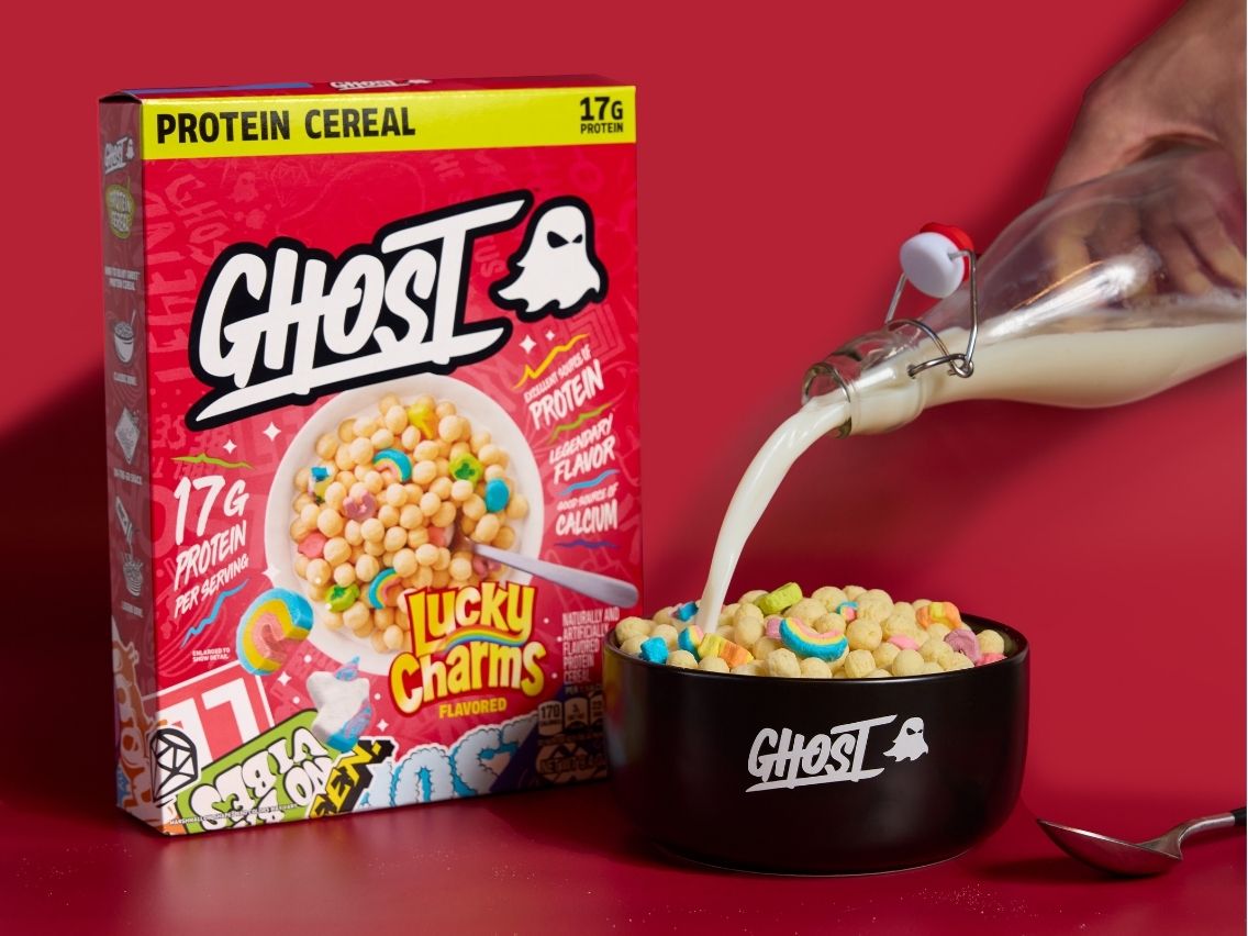 A box of GHOST® PROTEIN CEREAL x LUCKY CHARMS™​ next to a bowl labeled “GHOST” with the cereal in it and someone pouring milk into the bowl.